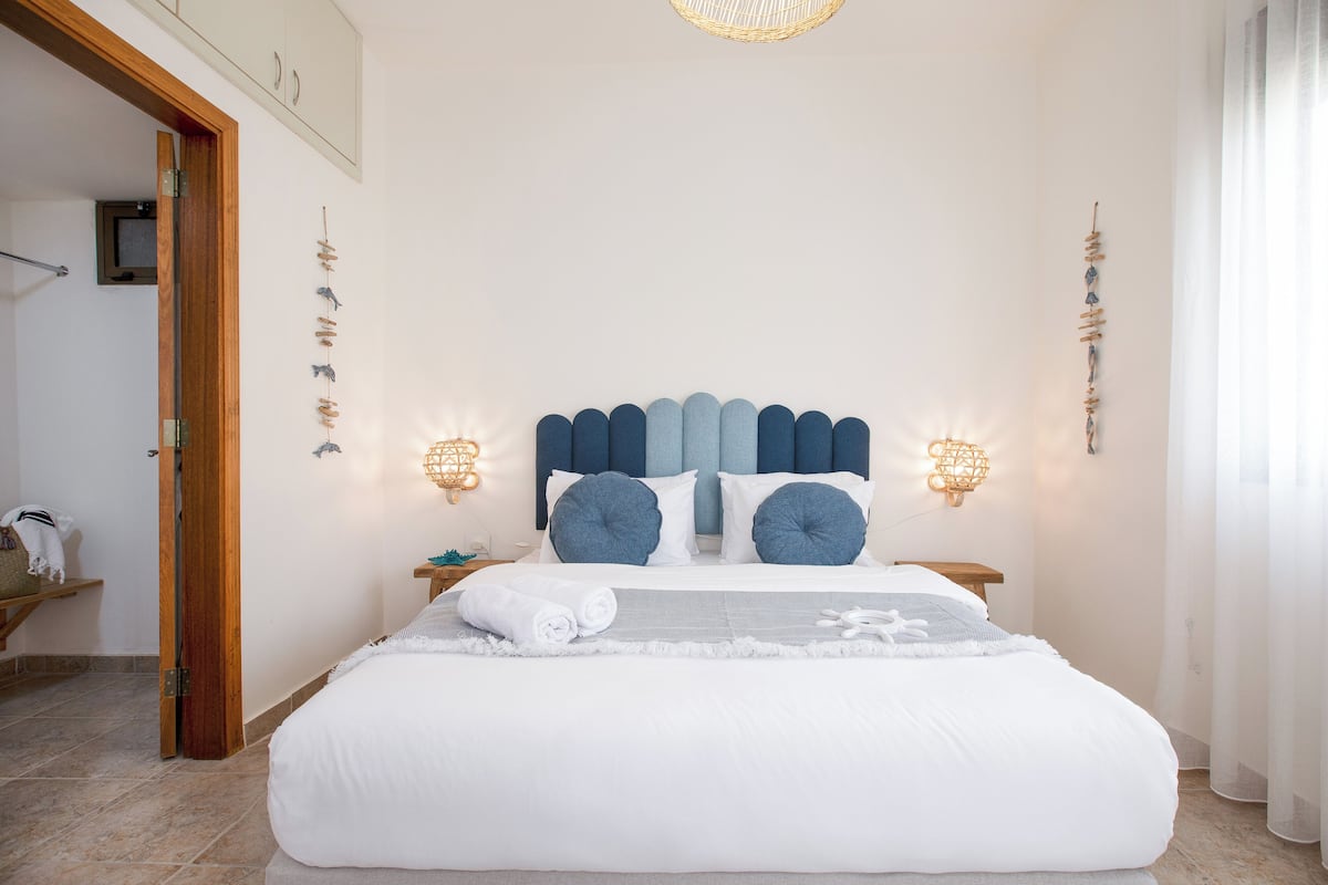 A neatly made bed with a white comforter and blue pillows, a bedside table with a lamp, and a wall-mounted light fixture.