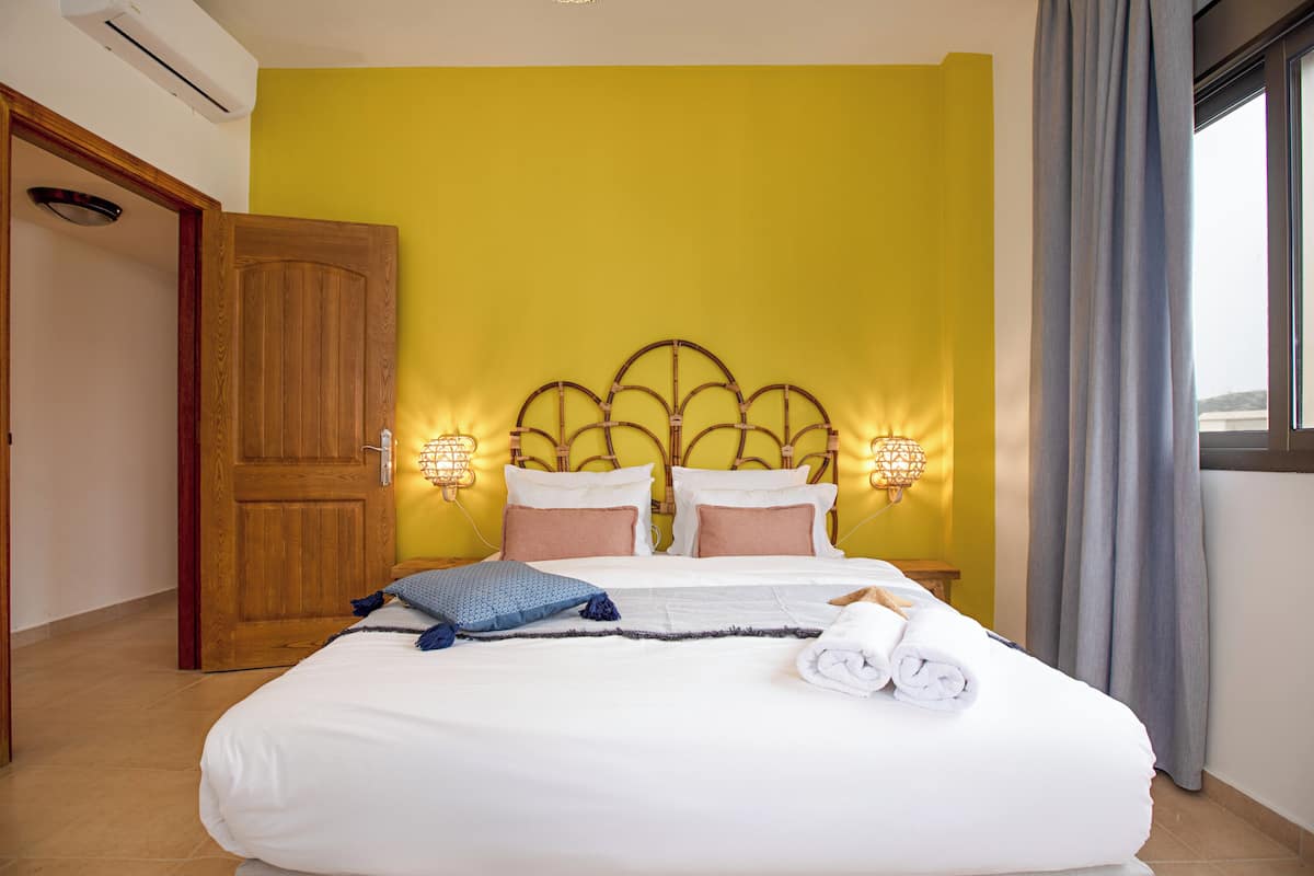 A hotel room with a wooden door, a bed with white linens, a headboard with integrated lighting, and a view of the outside through a window with curtains.