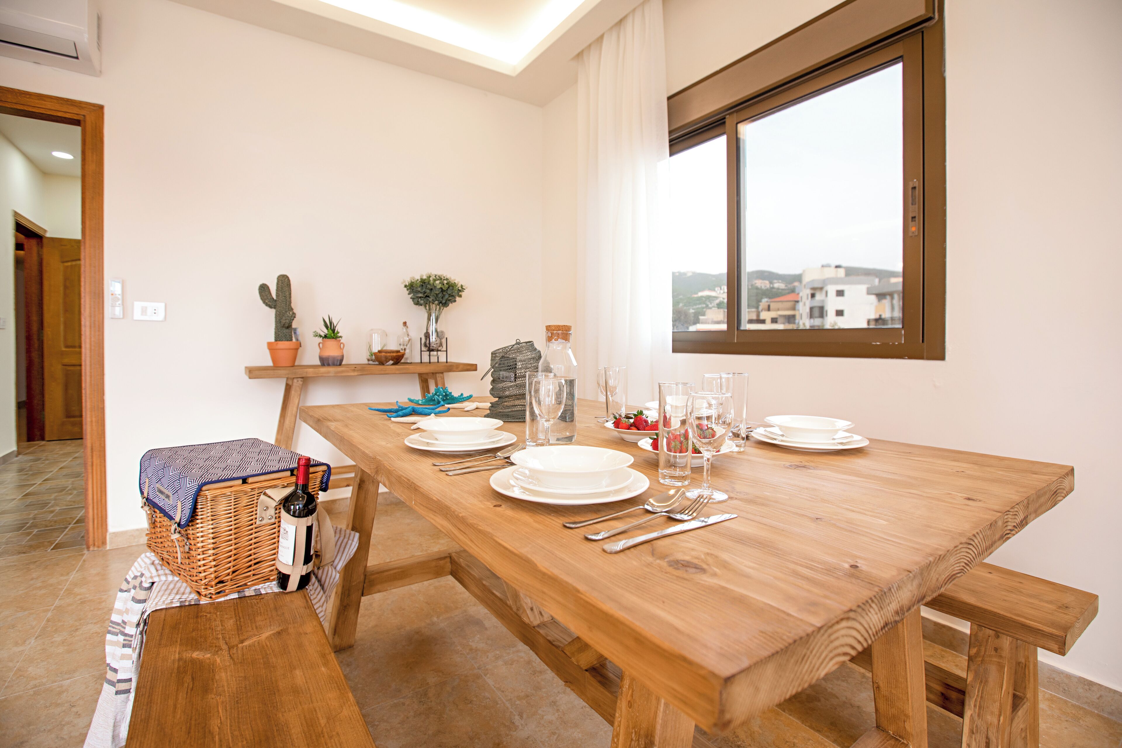 Uma área de refeições com uma mesa de madeira posta para quatro pessoas, incluindo pratos, copos e talheres. Há uma cesta de vime com uma garrafa e copos, uma pequena planta e uma janela com vista para prédios.