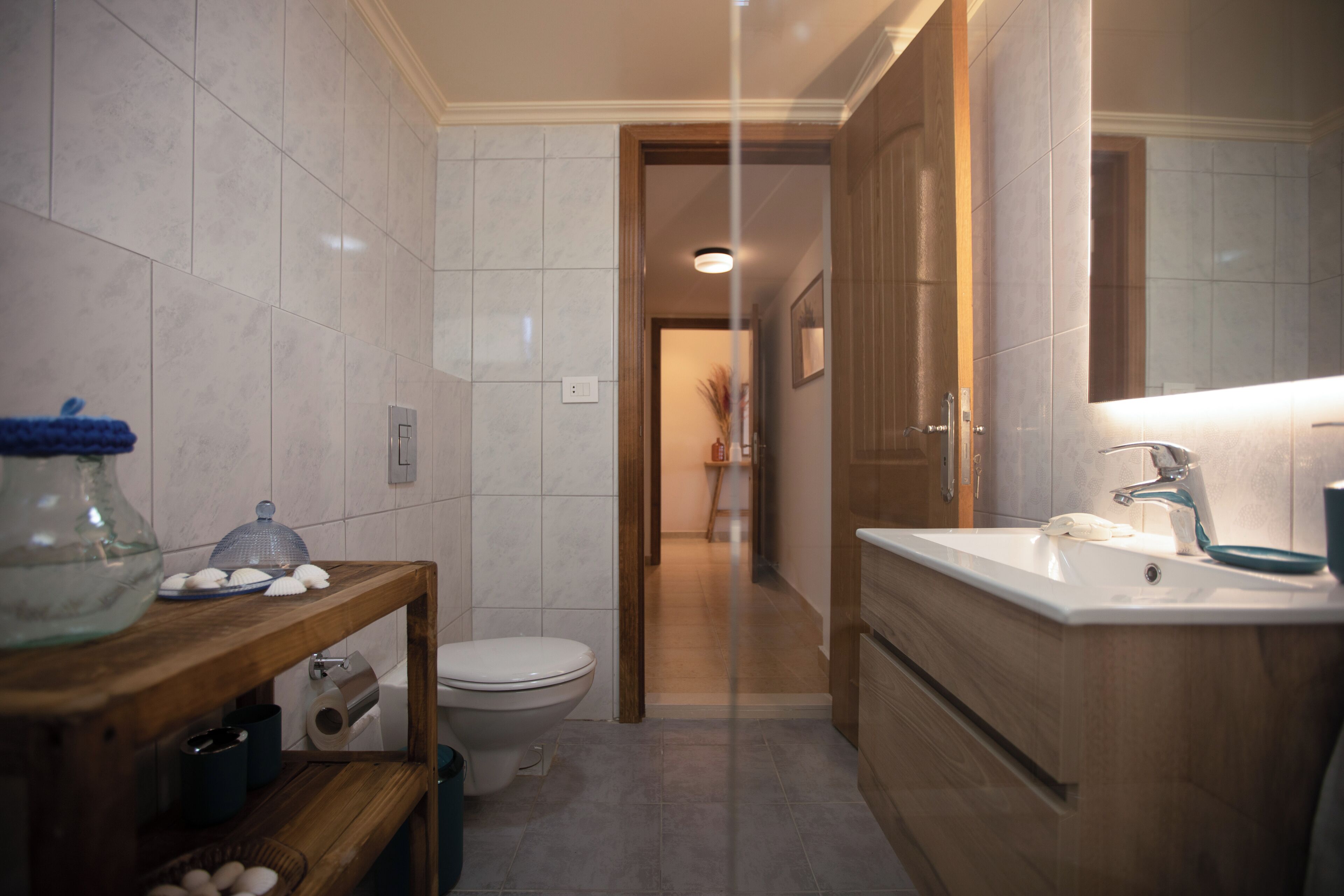 Une salle de bain avec des toilettes, un lavabo et un miroir. On y trouve une étagère avec un bocal et un panier d’œufs. Les murs sont recouverts de carreaux, et une porte en bois donne accès à une autre pièce.