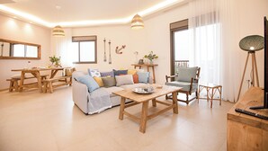 Amplia sala de estar con sofá de madera, una mesa de centro y una mesa de comedor. Hay dos lámparas colgantes, una lámpara de pie y un ventanal con cortinas.