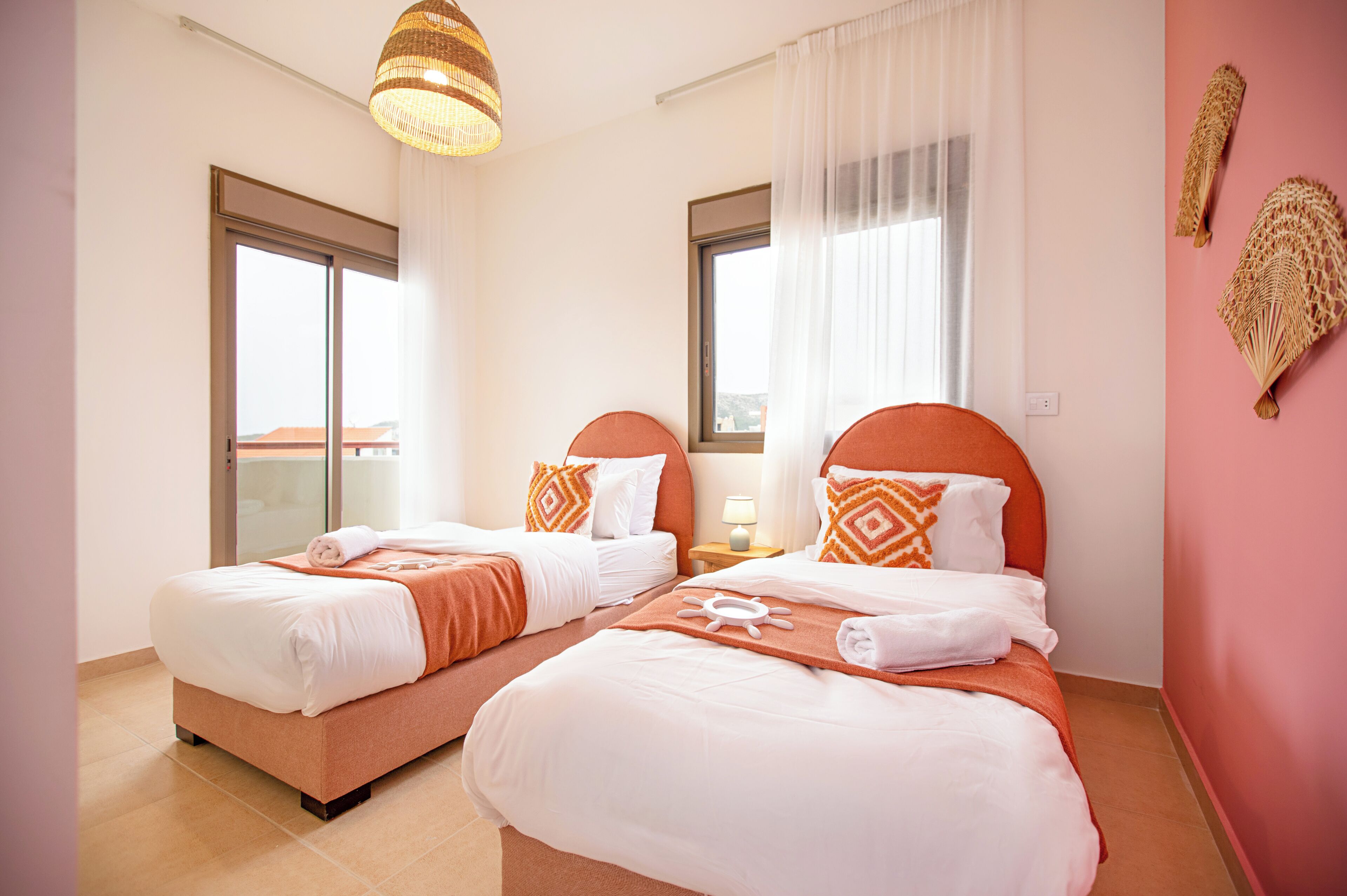 A hotel room with two beds, each with white and orange bedding, a large window, and a hanging light fixture.