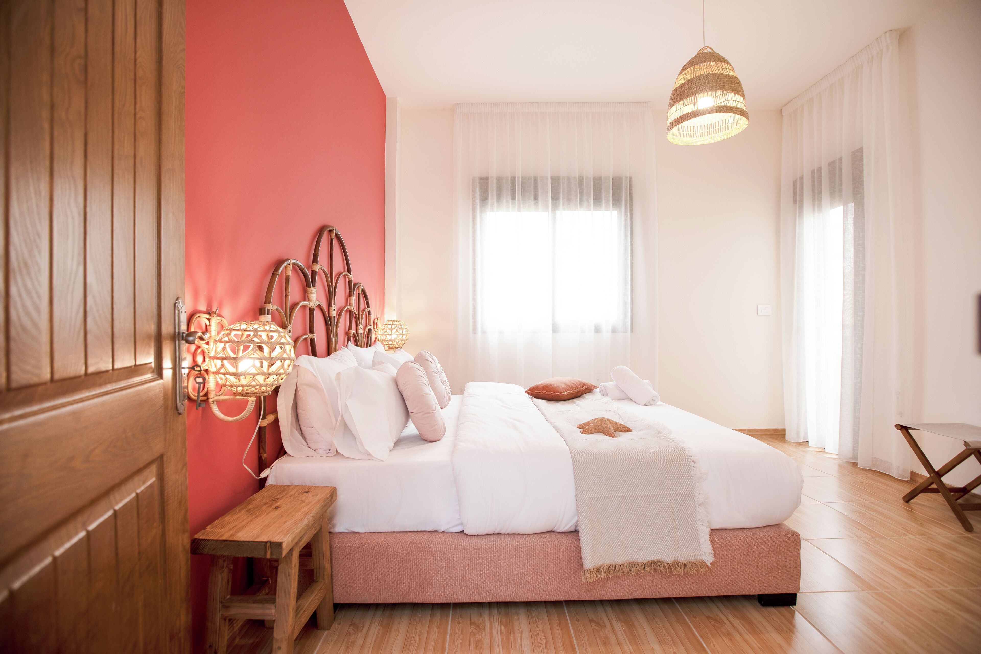 Een slaapkamer met een houten vloer, een bed met wit beddengoed, een nachtkastje, een hanglamp en een raam met dunne gordijnen.