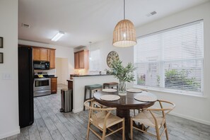 Una cocina moderna con armarios de madera, un refrigerador negro y un horno de acero inoxidable. Hay un comedor con una mesa redonda y sillas de mimbre.