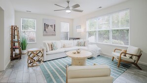A spacious living room with a white sofa, a round coffee table, and a wooden side table. There is a ceiling fan, large windows with blinds, and a striped rug on the floor.
