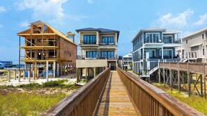 Une promenade en bois mène à une propriété située en bord de mer, qui comprend plusieurs bâtiments, dont une maison en construction et un immeuble à deux étages terminé, doté de balcons.