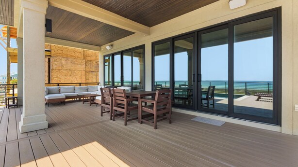 Un espace extérieur abrité doté d’une terrasse en bois, d’un coin salon avec un canapé et des fauteuils, et de grandes portes-fenêtres coulissantes offrant une vue sur l’océan.