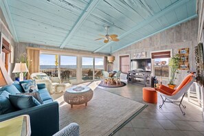 A spacious living area with a large window overlooking the ocean, a ceiling fan, and a dining area with chairs and a table. - Gulf Front Beach House | Spacious Deck | Outdoor Shower | Sleeps 10 (Gulf Shores)