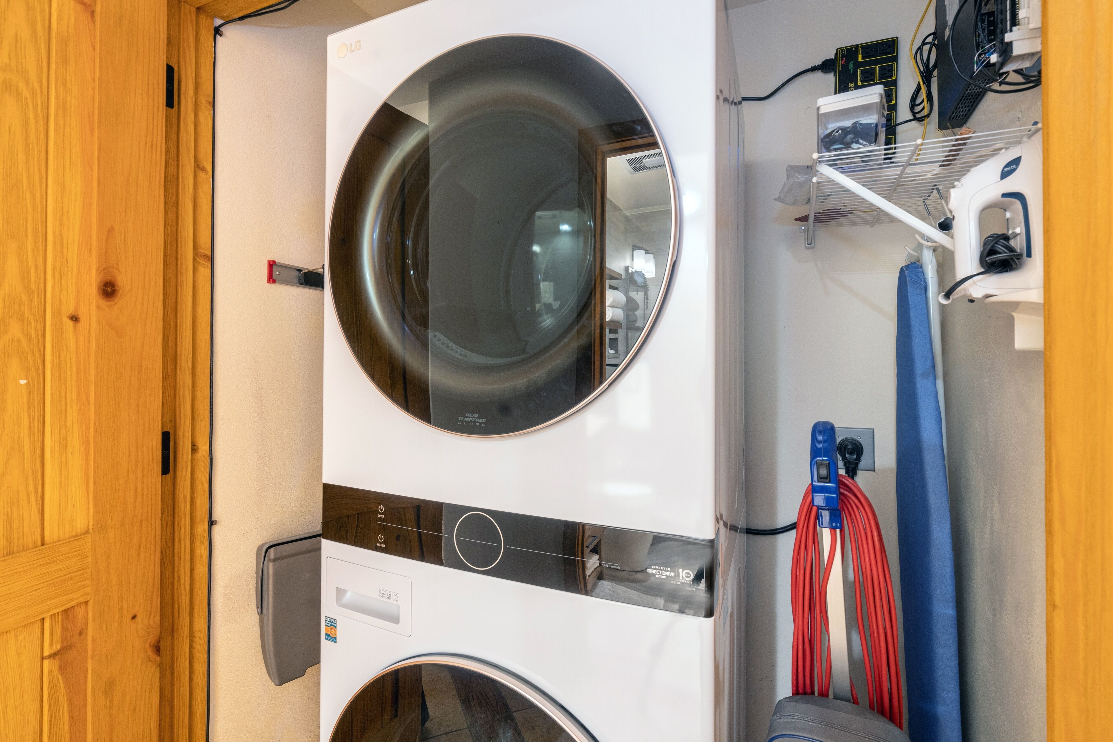 A front-loading washing machine and dryer in a laundry room with a wooden door, a shelf with a camera, and a blue towel hanging.