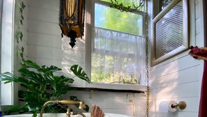 Una bañera con grifo dorado, rodeada de follaje y una ventana con vistas a árboles.