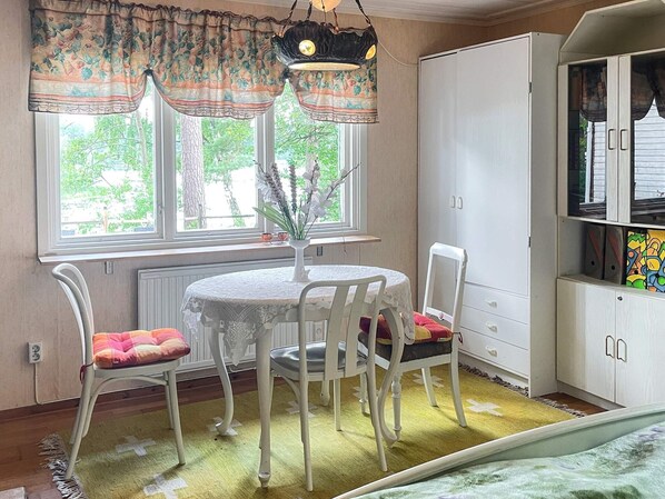 A room with a round table, two chairs, a radiator, a window with floral curtains, and a cabinet with colorful artwork.