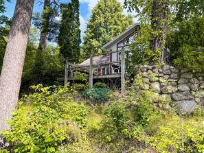 A rustic cabin with a wooden deck, stone wall, and lush greenery.