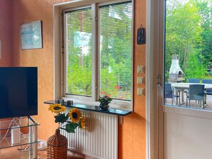 A room with a large window, a television, a wall-mounted shelf with a vase of sunflowers, and a view of a garden and outdoor seating area.