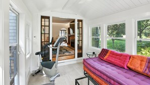 Habitación con cinta de correr, sofá morado con cojines estampados y ventanas amplias que dan a vistas de vegetación.