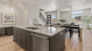 A modern kitchen with a large island, a dining area with a table and chairs, and a staircase leading to another level. - The Crash Pad 5 Bedroom Townhouse (St. George)