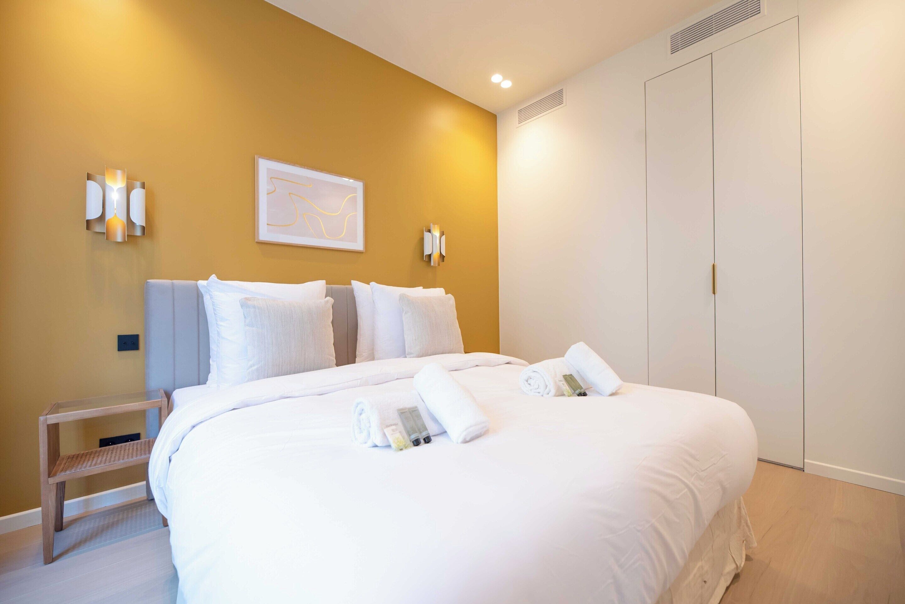 A neatly made bed with white linens and towels, a bedside table with a lamp, and a wall-mounted artwork.