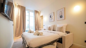 A hotel room with a bed, a TV mounted on the wall, and a window with curtains. - Elegant Apt With AC - 2br-6p -palais Garnier (Paris)