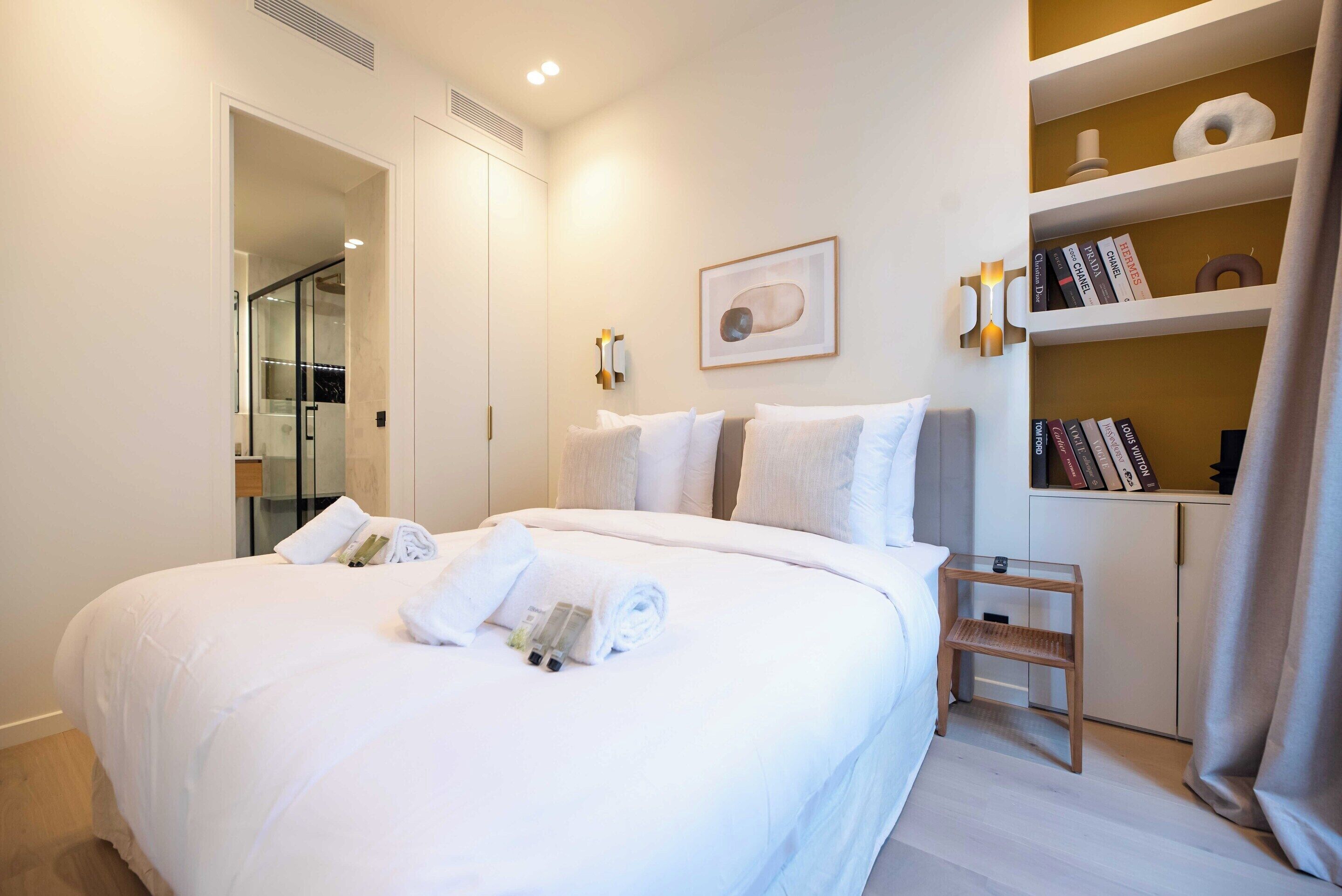 A neatly made bed with white linens and towels, a bedside table with a lamp, and a bookshelf with books and decorative items.