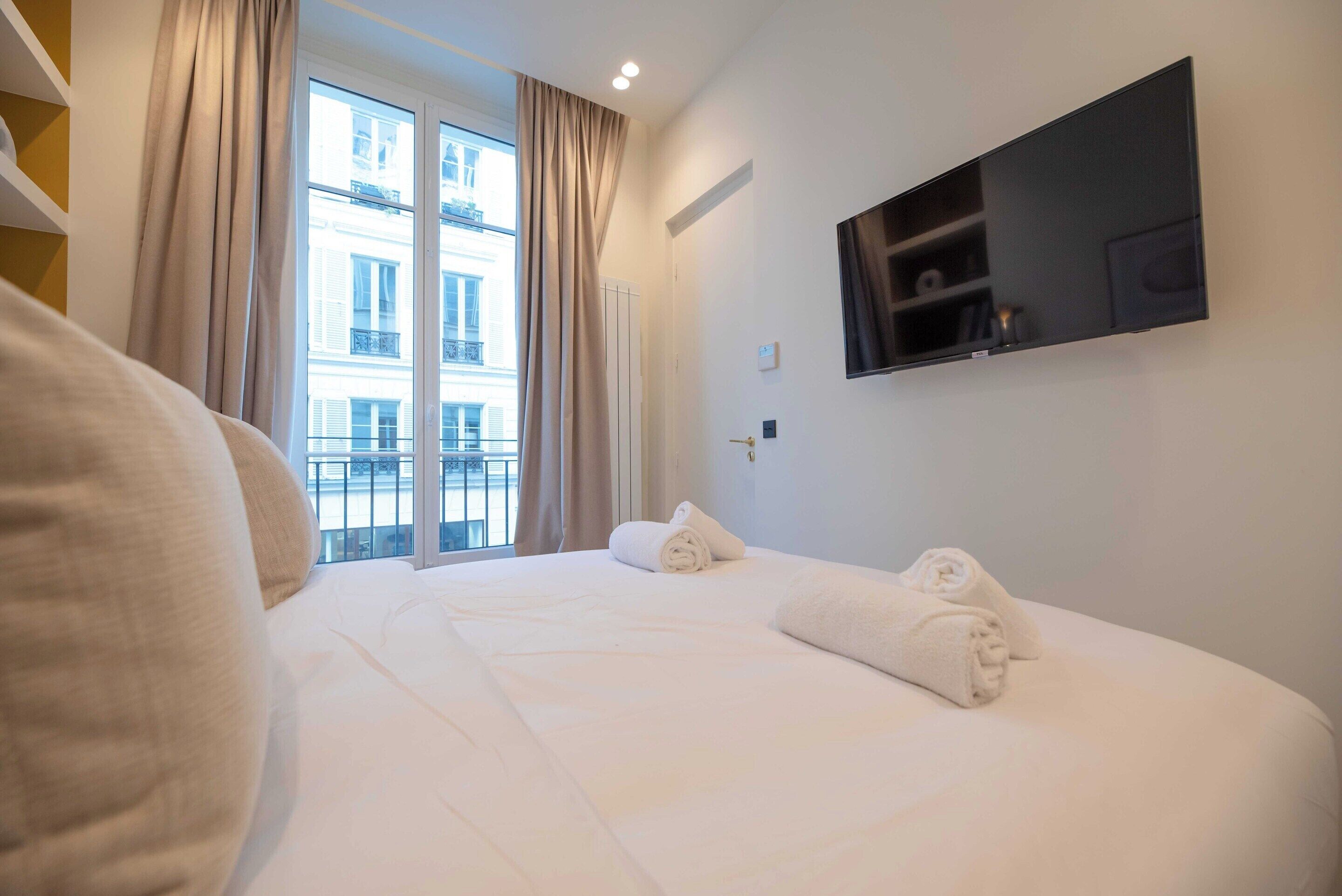 Una habitación de hotel con una cama grande, un televisor montado en la pared y una ventana con cortinas.