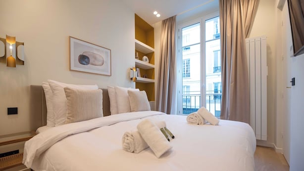 A hotel room with a large bed, a bedside table, a wall-mounted shelf with books, a framed picture, and a window with curtains. - Elegant Apt With AC - 2br-6p -palais Garnier (Paris)