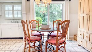 A dining area with a round glass table surrounded by four wooden chairs with red cushions, a chandelier, and a view of greenery through a window.