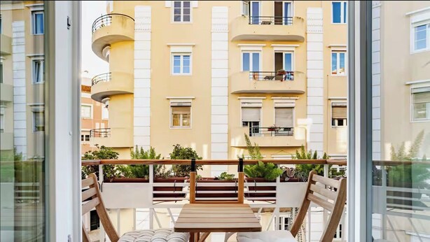 Un balcon doté de mobilier en bois, comprenant une table et deux chaises, donnant sur un immeuble d’habitation avec des balcons et des fenêtres.
