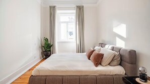 Une chambre à coucher moderne avec un grand lit, une plante en pot et une vue sur la ville par la fenêtre.