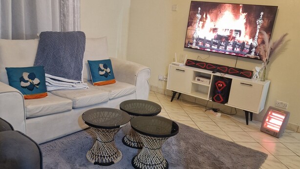 A living room with a sofa, two black coffee tables, a TV displaying a fire scene, a white cabinet, and a heater on the floor. - Double K Loft 1-bedroom apartment in Nairobi (Nairobi)