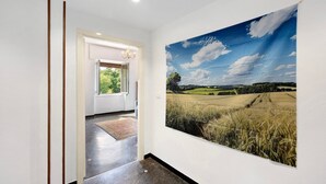 Un couloir orné d’un grand tableau représentant un paysage, une porte donnant sur une pièce et un lustre.