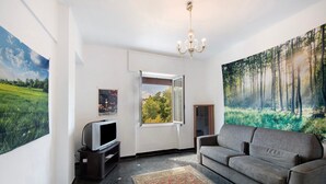 Un salon avec un canapé, un téléviseur posé sur un meuble, un grand tableau représentant une forêt et une fenêtre.