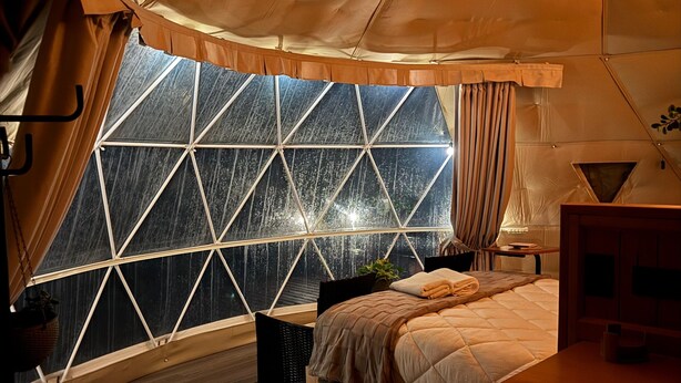 Ein zeltartiges Zimmer mit großem Fenster, einem Bett, einem Schreibtisch und einer Hängeleuchte.