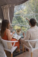 Una tienda de campaña con un ventanal grande, dos personas sentadas en una mesa con un libro y una planta, y vistas a árboles desde el exterior.
