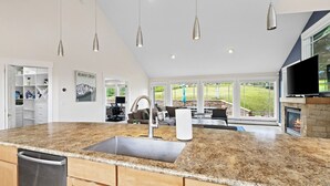A modern kitchen with granite countertops, a stainless steel sink, and wooden cabinets. There is a dining area with a fireplace and a large window overlooking a garden.