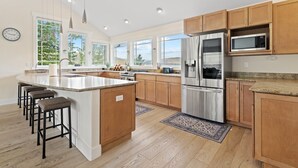 A modern kitchen with a central island, stainless steel appliances, wooden cabinets, and large windows offering views of the outdoors.
