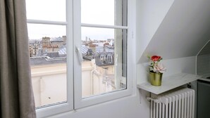 A room with a window overlooking a cityscape, a radiator, and a small shelf with a vase of flowers.