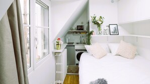 A compact bedroom with a bed, a radiator, a small kitchenette, and a window with curtains.