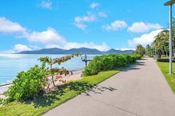 A paved walkway alongside a body of water with distant mountains, a clear sky, and lush greenery.