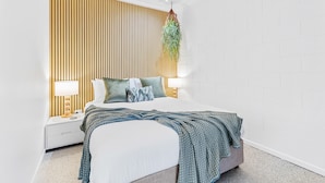 A modern bedroom with a bed, bedside tables, a hanging plant, and a wooden slat wall. - Rainforest Reef Retreat - Esplanade Ground-floor Apartment (Cairns)