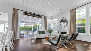 Un salon spacieux avec un canapé blanc, une table basse ronde et deux fauteuils en cuir noir. On y trouve une cheminée surmontée d’un miroir, ainsi que de grandes fenêtres avec des rideaux.