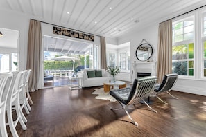 Een ruime woonkamer met een witte bank, een ronde salontafel en twee zwarte leren fauteuils. Er is een open haard met een spiegel erboven, en grote ramen met gordijnen.