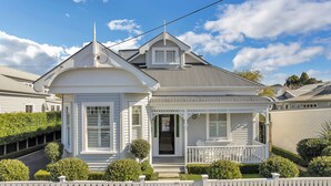 Une maison de style victorien à deux étages, dotée d’une véranda qui fait le tour de la maison, d’une entrée avec une porte et d’un petit balcon.