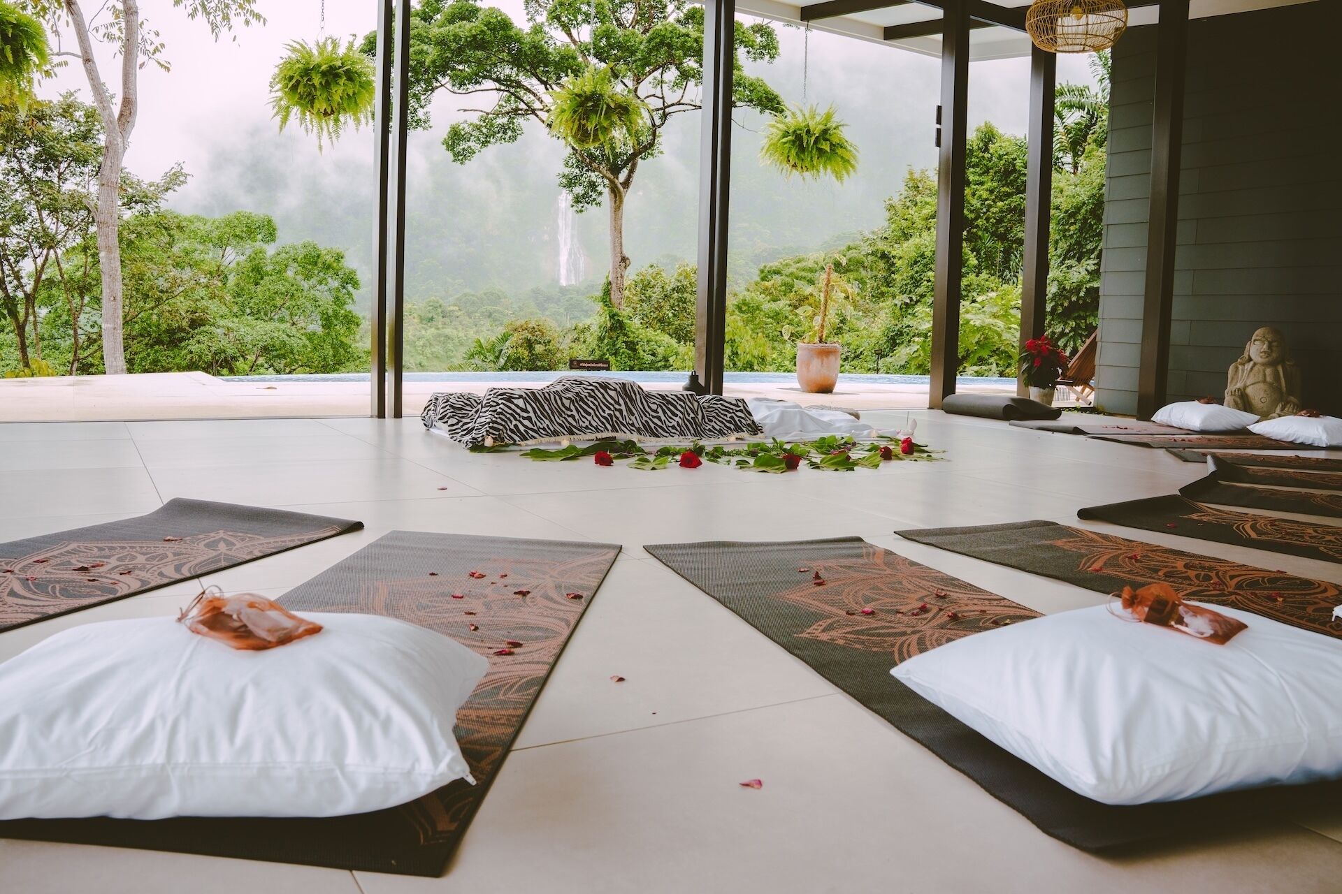 Un espace intérieur paisible, offrant une vue sur une végétation luxuriante et une cascade. On y trouve un espace dédié à la méditation, avec des tapis et des coussins, un coin salon agrémenté d’un coussin à motifs zébrés, et une statue de Bouddha.