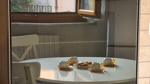 A round table with four teacups and a plate of cookies, a white chair, and a window with a view of a building and a fence. - Verdi HOUSE - Relaxation and Flavors (Savigliano)