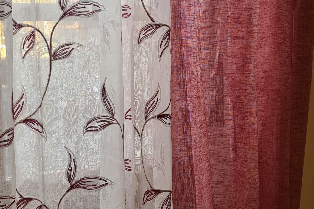 A room with a window featuring sheer curtains with a leaf pattern and a solid red curtain.