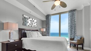 A bedroom with a large bed, a nightstand, a lamp, a ceiling fan, and a view of the ocean.