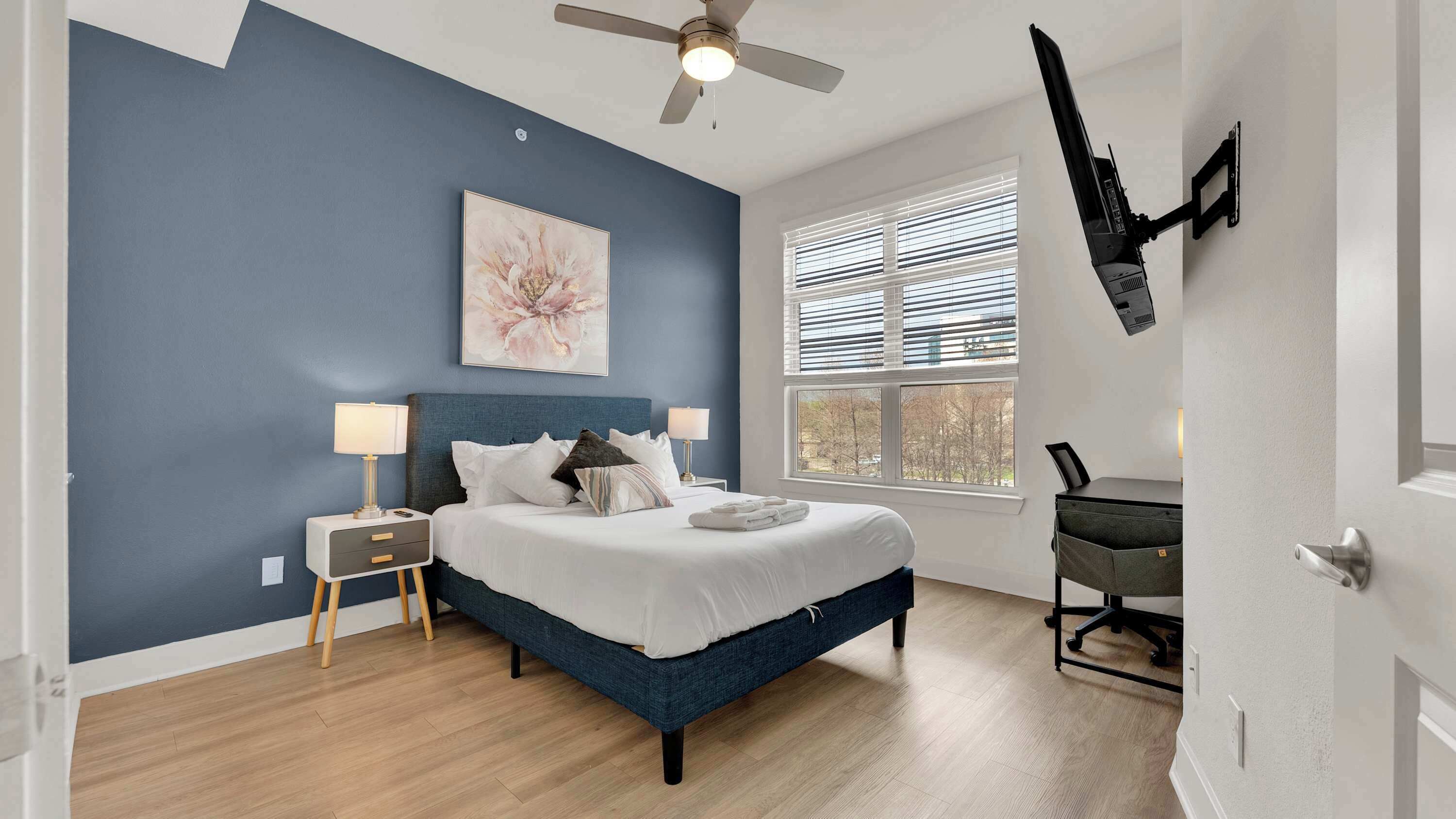 Une chambre à coucher comprenant un lit, des tables de chevet, un ventilateur de plafond, une fenêtre avec des stores et un téléviseur fixé au mur.