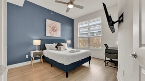 Une chambre à coucher comprenant un lit, des tables de chevet, un ventilateur de plafond, une fenêtre avec des stores et un téléviseur fixé au mur.