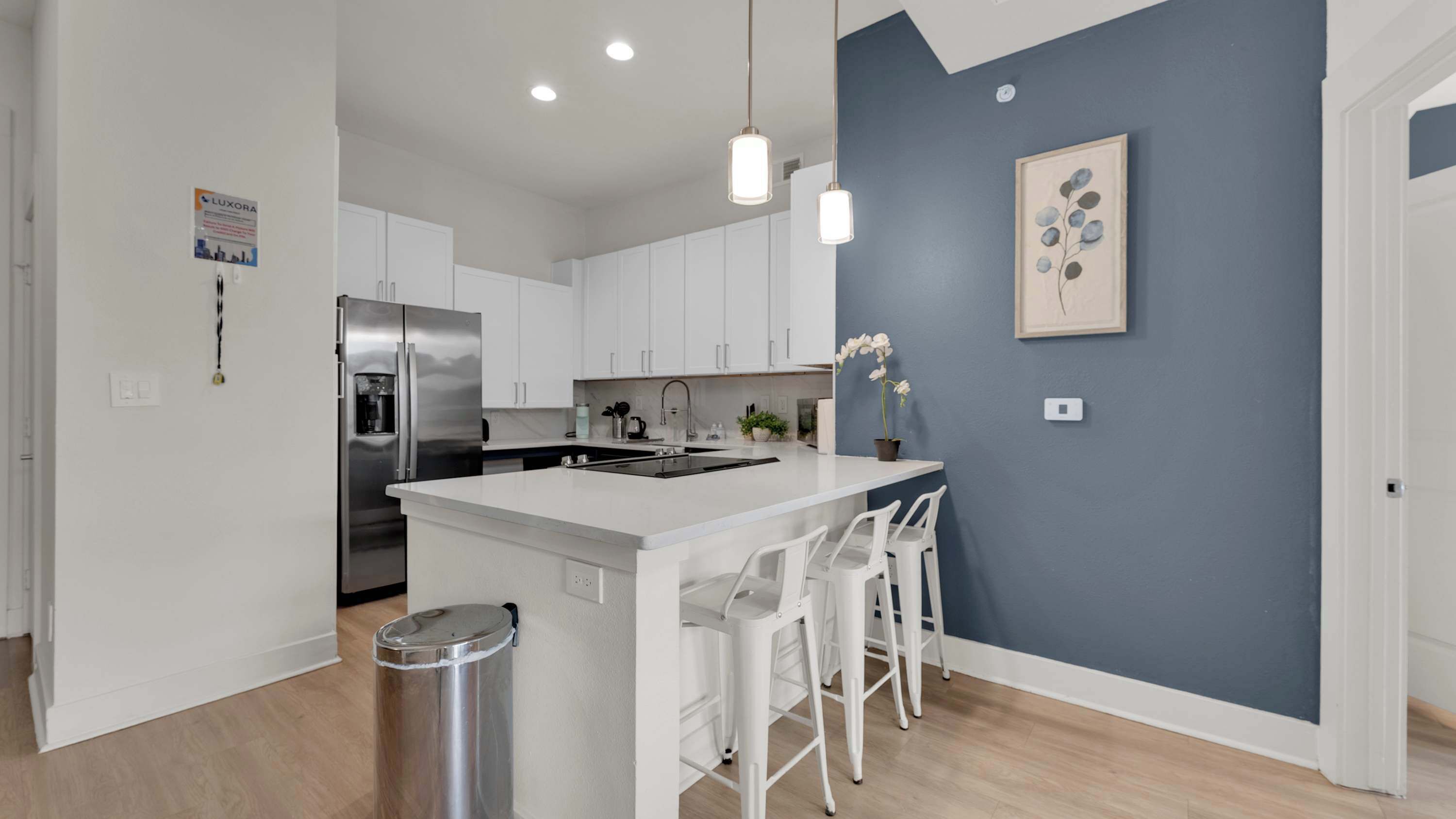 A modern kitchen with white cabinets, a stainless steel refrigerator, a sink, and a bar area with white stools.