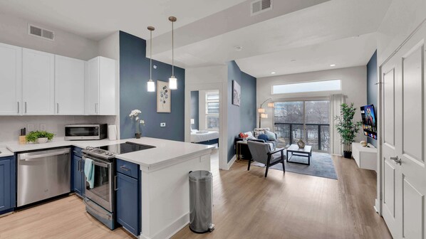 A modern kitchen with a central island, a dining area with a sofa and coffee table, and a balcony with a TV.
