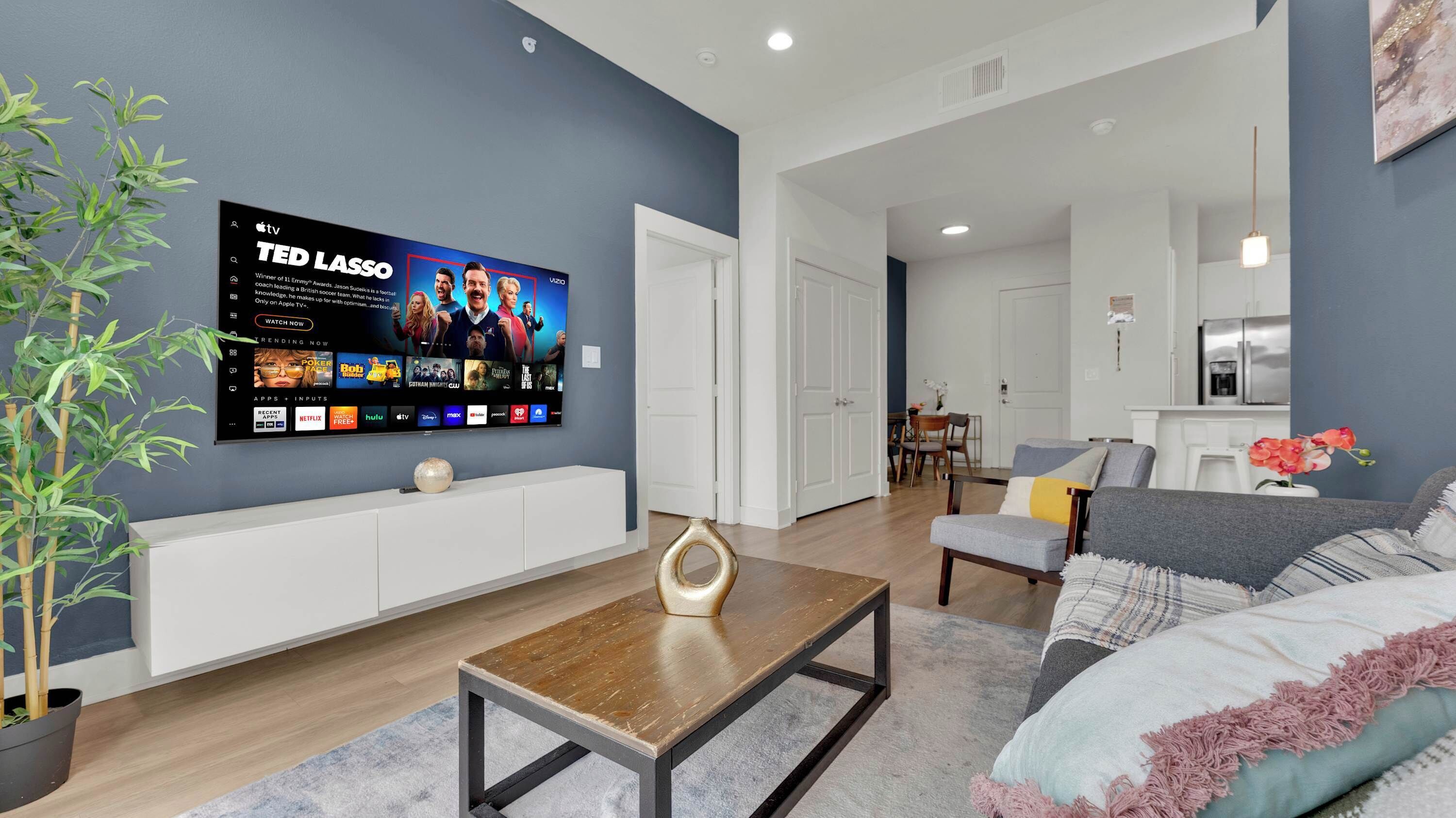 Ein modernes Wohnzimmer mit Flachbildfernseher, der eine Fernsehsendung zeigt, einem Couchtisch, einer Couch und einem Essbereich im Hintergrund.